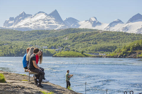 Bildet viser fire ungdommer som sitter på en benk langs vannet, med utsikt over Saltstraumen. I bakgrunnen står en mann og fisker med fiskestang og vi ser de snødekte Børvasstindan i bakgrunnen. 