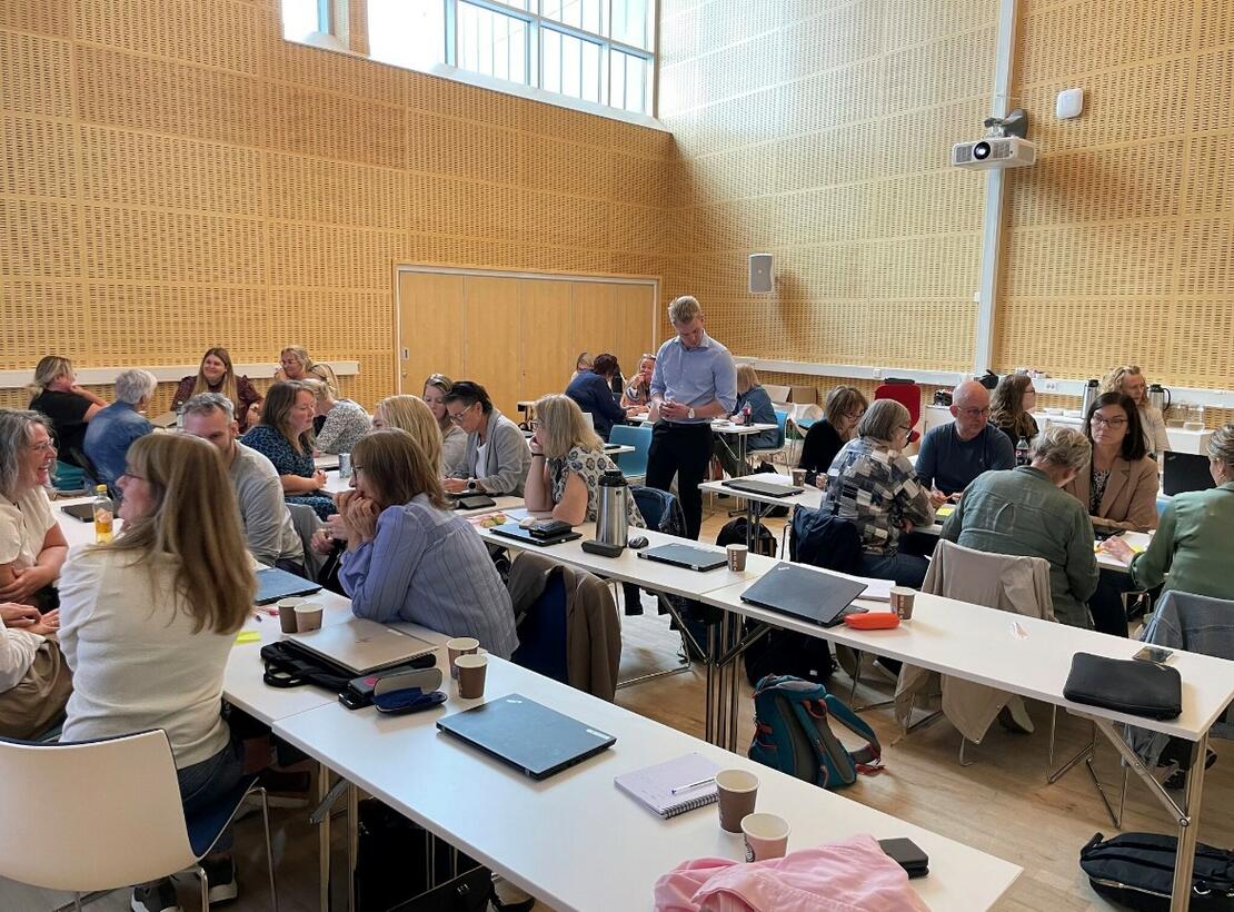 Bilde som viser flere personer som deltar på en workshop, personer som sitter og jobber og diskuterer ved flere bord. 