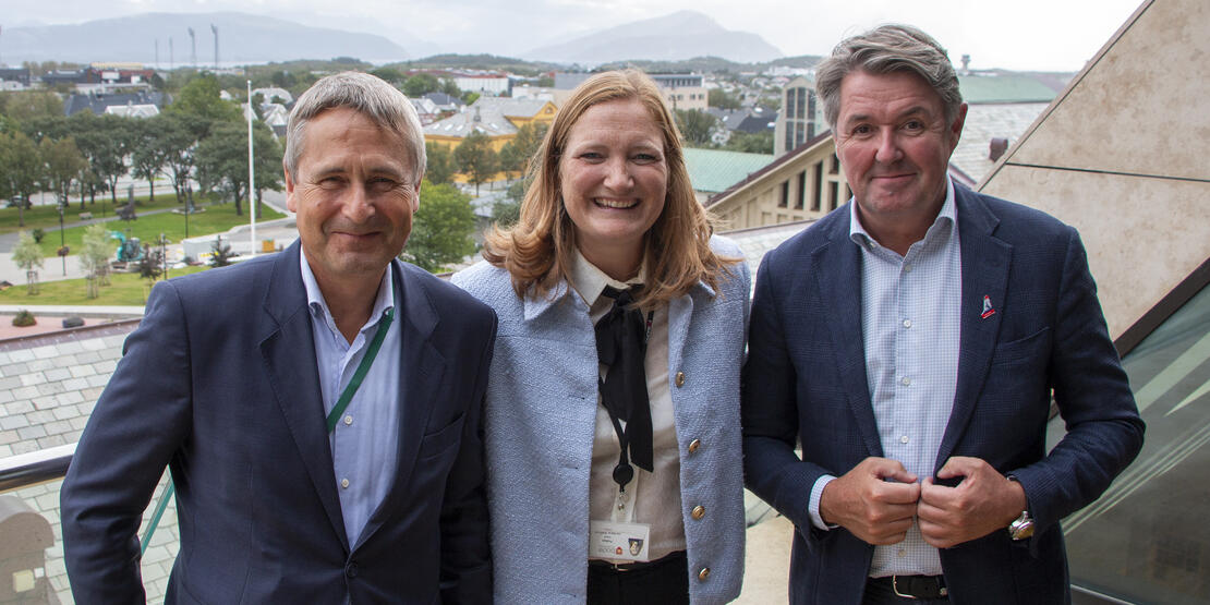 Tre personer som smiler. Utsikt over deler av rådhusparken og deler Bodø sentrum, fjell og skyer. 