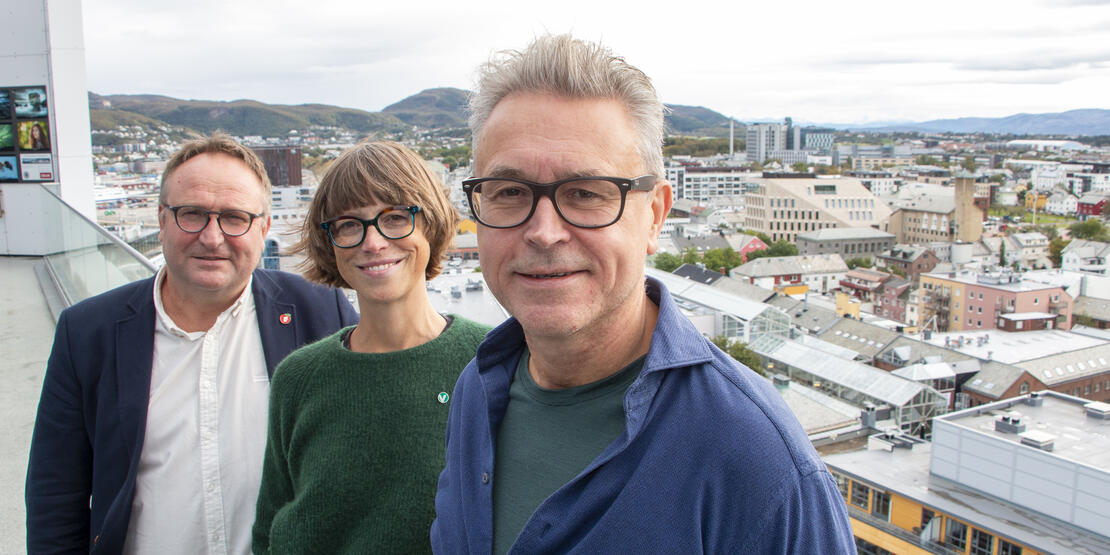 Tre personer som smiler. Bildet er tatt fra balkongen i øverste etasje på Scandic Havet, og viser deler av Bodø sentrum i bakgrunnen, fjell og himmel. 