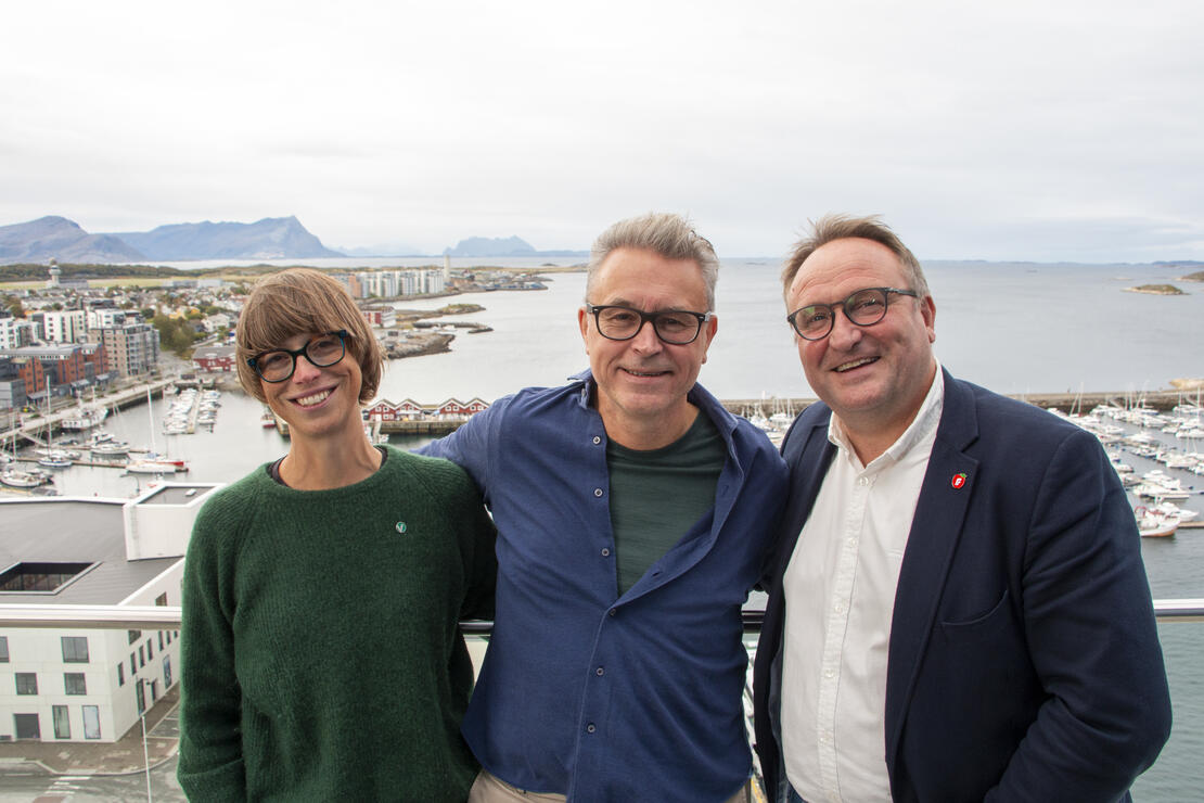 Tre personer som smiler. Bildet er tatt fra balkongen i øverste etasje på Scandic Havet, og viser deler av Bodø sentrum, hav, fjell og himmel. i bakgrunnen. 
