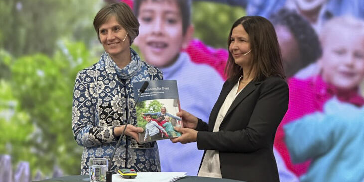 Bildet viser Mari Rege som gir rapporten, i papirformat, til Kjersti Toppe. I bakgrunnen vises det samme forsidebildet i større format. Bildet viser barn i aktivitet ute. 