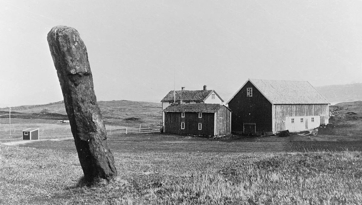 Gammelt sort/hvitt-bilde som viser en bauta-stein ved Rostadgården på Hernes i Bodø. Gress, himmel,det er tre ulike bygg på gården. 