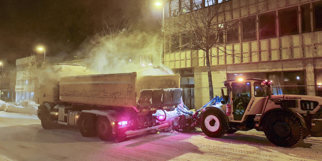 Bilde av lastebil og brøytebil, snø, gate, gatelys, bygninger. 