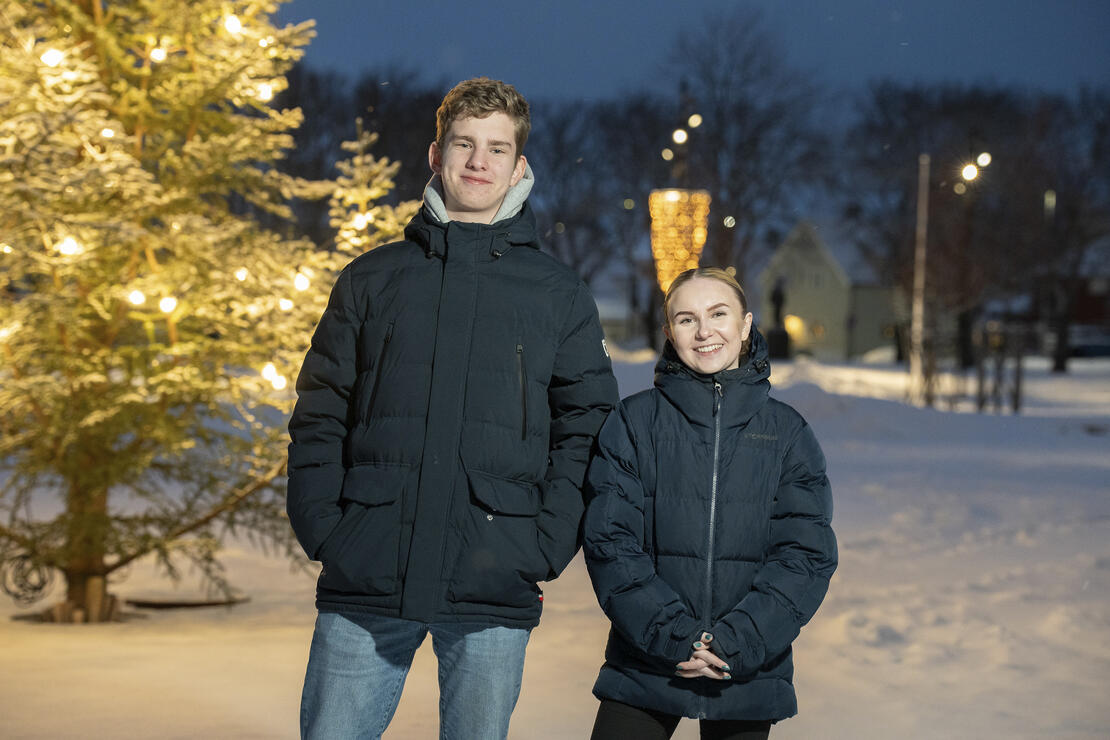 Bildet viser to personer som smiler. Deler av rådhusplassen vises i bakgrunnen. Snø, trær, hus, juletre og vinterlys. 