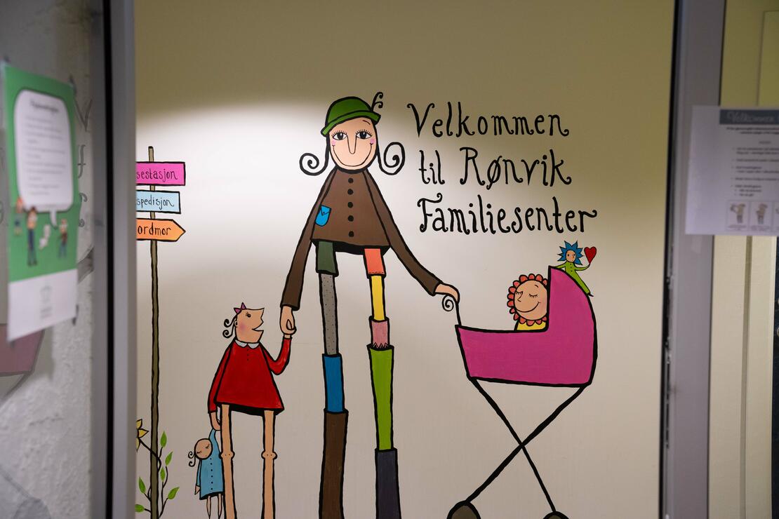 Bilde av veggen og veggmaleri med tegnede figurer som framstiller en familie på tre, og en tekst der det står "Velkommen til Rønvik Familiesenter"