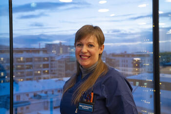 Person i sykepleieruniform fotografert oppe foran et vindu med utsikt over deler av Bodø sentrum. 