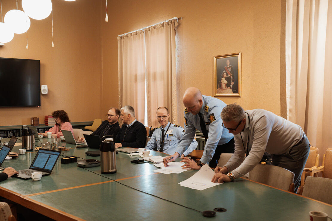 To personer, hvorav en av dem er i politiuniform), står til høyre i bildet og signerer en avtale ved et langbord. Innover i bildet, på samme langbord, sitter fire synlige personer og observerer signeringen. 