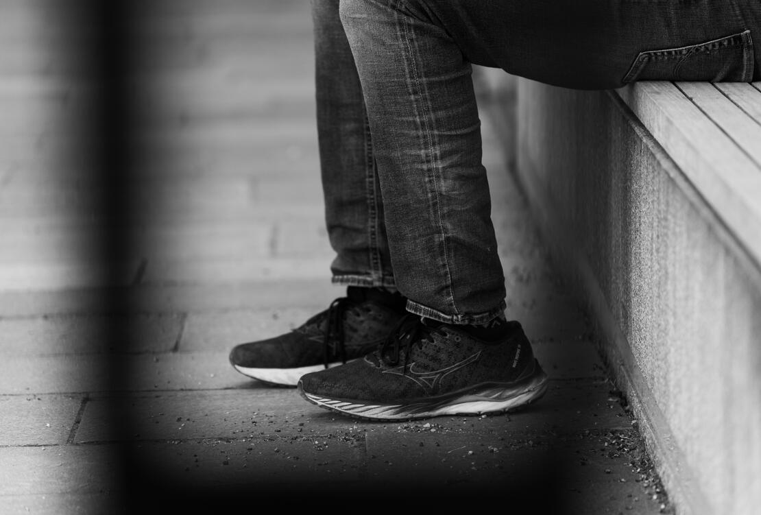 Bildet er sort og hvitt. Det er tett på et par ben. Dongeribukse og svarte sko og hvite såler, og personen ser ut til å sitte på en benk. 
