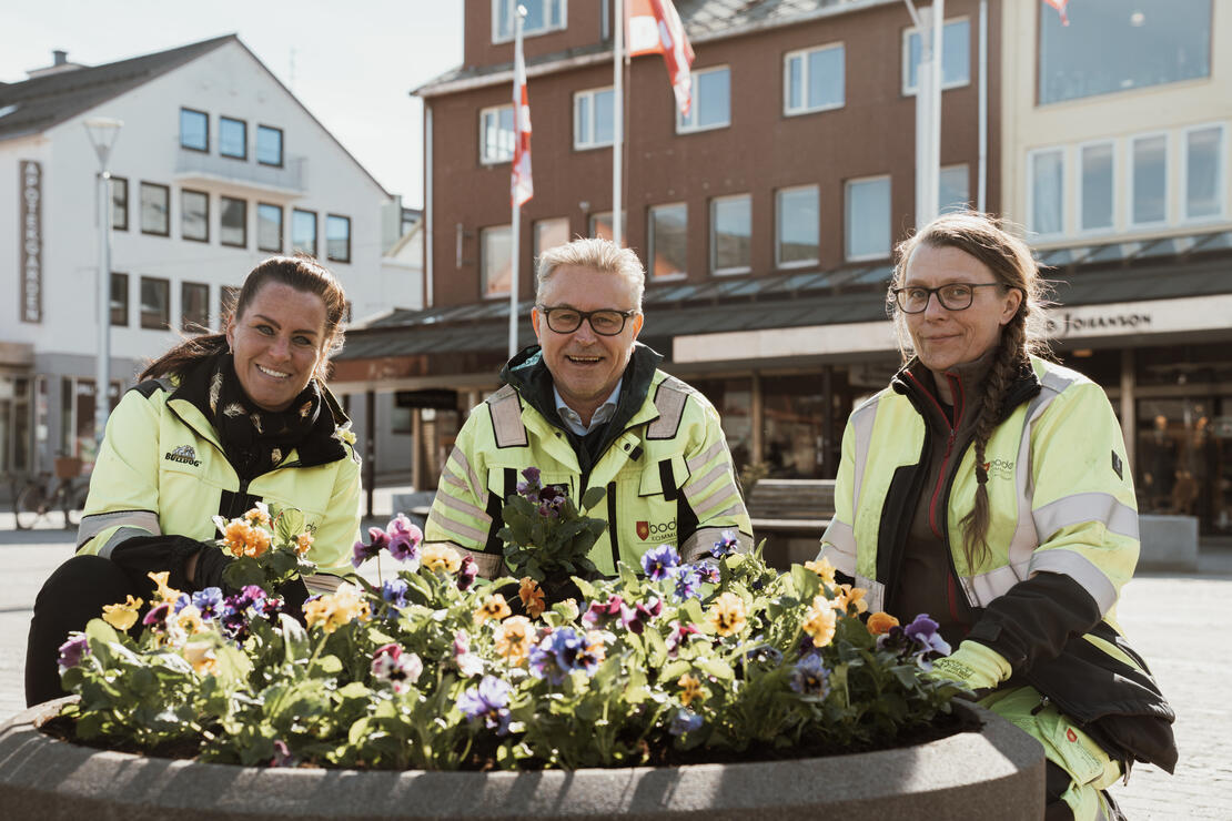 tre personer, to kvinner og en mann, i gule arbeidsjakker sitter på huk foran en blomsterkasse med mange blomster i forskjellige farger, på et åpent uteområde i en by. De ser rett i kamera og smiler. 