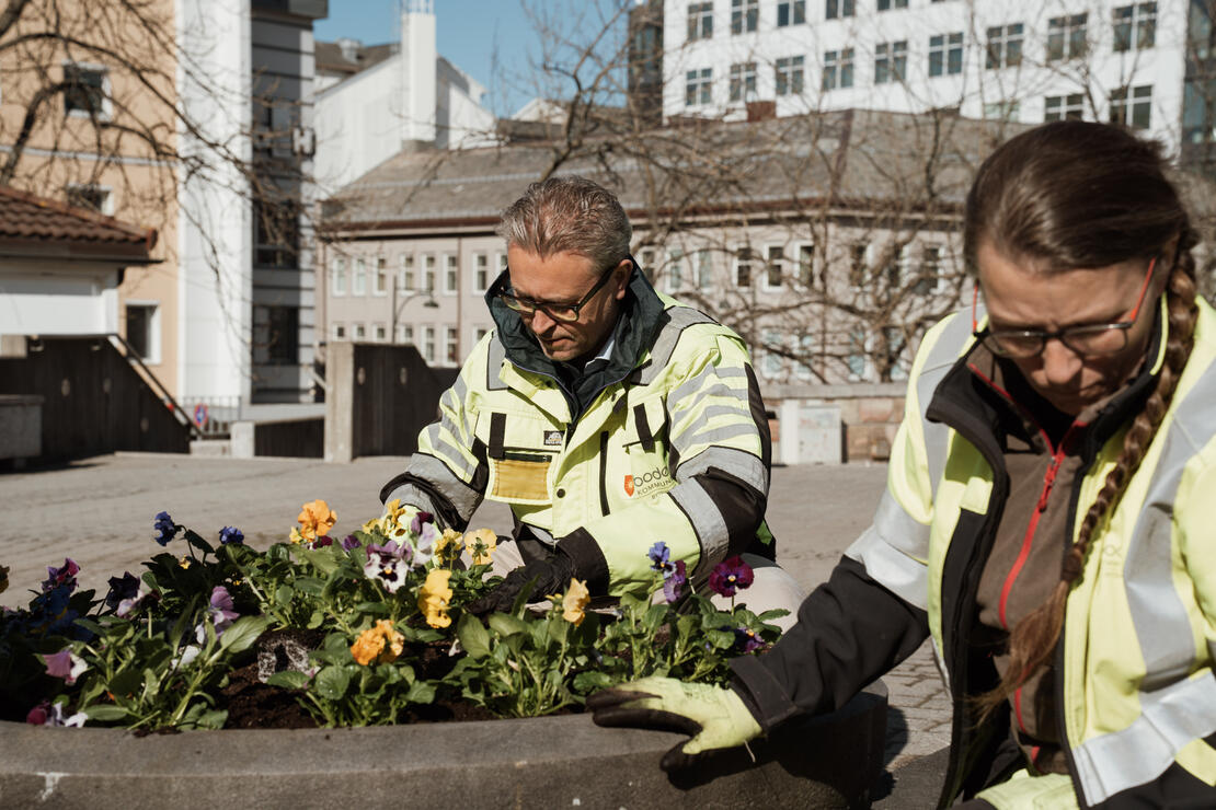 To personer, en mann og en kvinne, sitter på huk på et åpent uteområde i et sentrumslignende uteområde. Begge sitter å jobber med å plante blomster i en rund stor blomsterkasse.