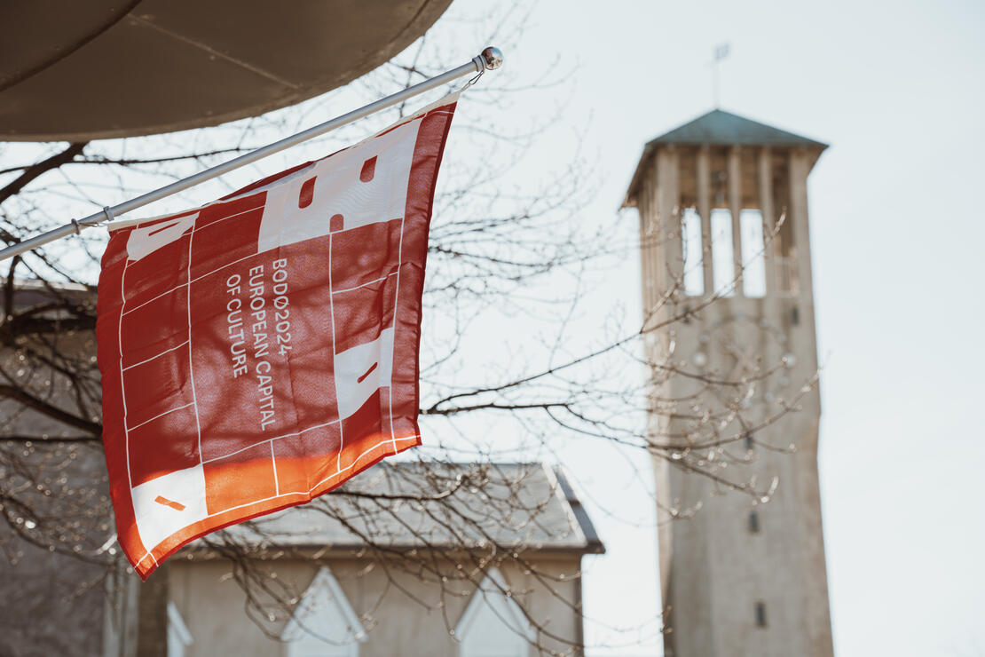 Et rødt flagg henger under et lite utspirng. Flagget har skriften Bodø2024 og i bakgrunnen kan et stort murhus og et klokketårn synes.