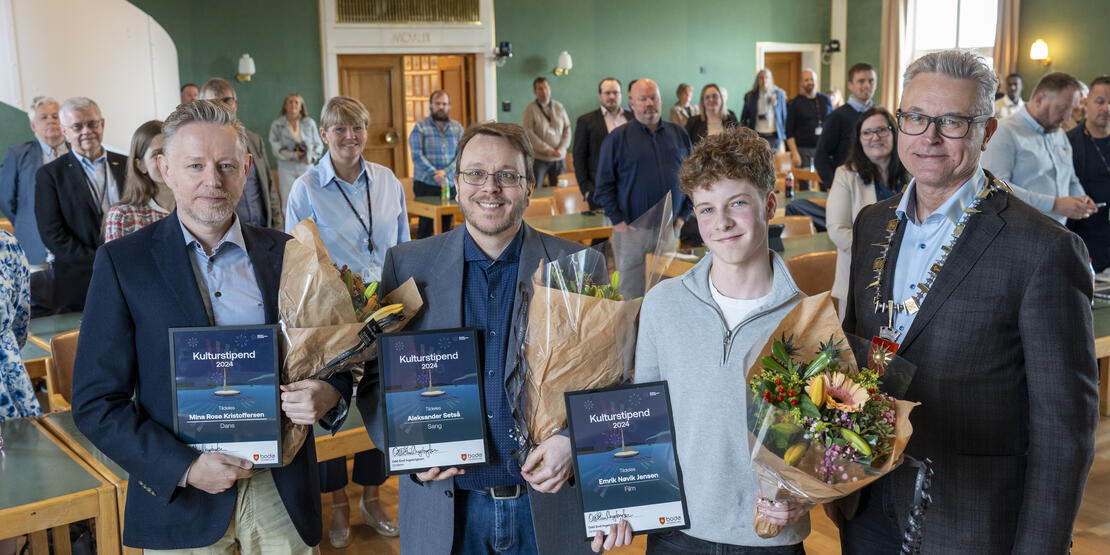 Bildet viser fire personer som smiler og står foran en sal med flere personer som vises i bakgrunnen. Tre av personene i forgrunnen holder et diplom og en blomsterbukett hver. Til høyre i bildet står ordføreren med ordførerkjede. 