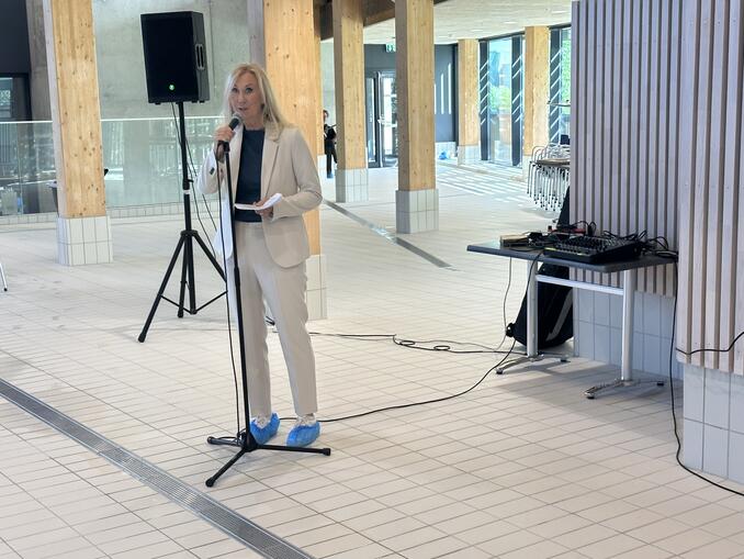Brita Bjørnbakk står bak en mikrofon. Det er gulv med fliser og vegger med tre, Det er glass bak også.
