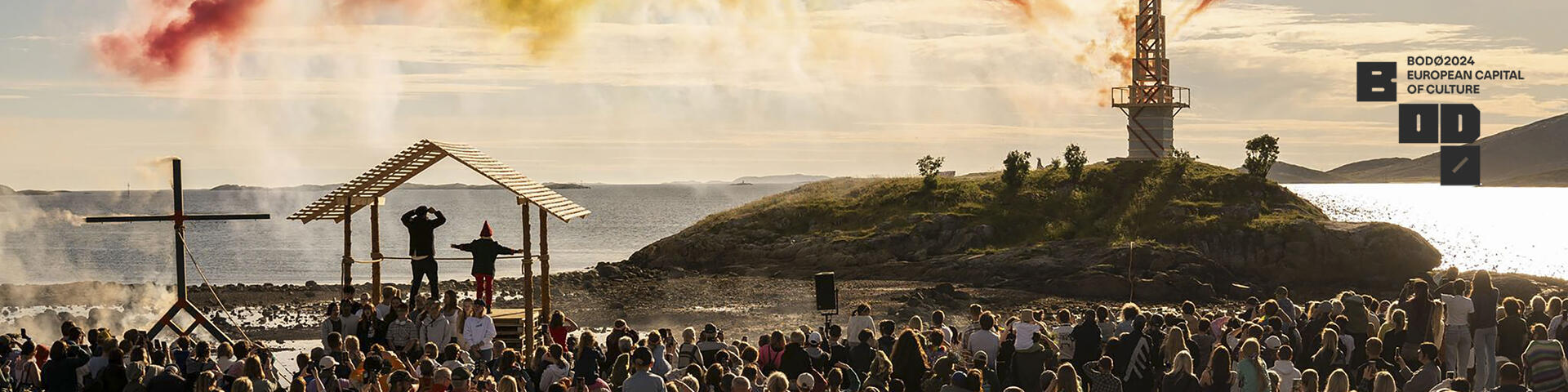 Bilde fra folkefest i Breivika i Bodø. Mange mennesker som ser på og fyrverkeri/farger. Hav og himmel. Bodø2024 sin logo er satt inn til høyre i bildet. Bildet lenker videre til Bodø2024s nettside. 