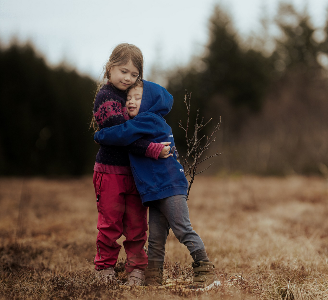 To barn, en jente og en gutt, holder rundt hverandre på en eng av gress. I bakgrunnen skimtes det mørke trær og en lys himmel.
