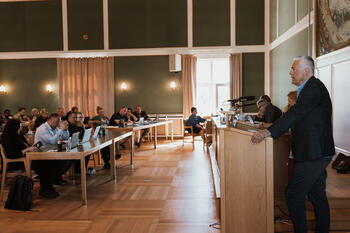 Kommunedirektøren står på talerstolen i bystyresalen i Bodø. Du ser medlemmer i bystyret som sitter ved bord i salen.