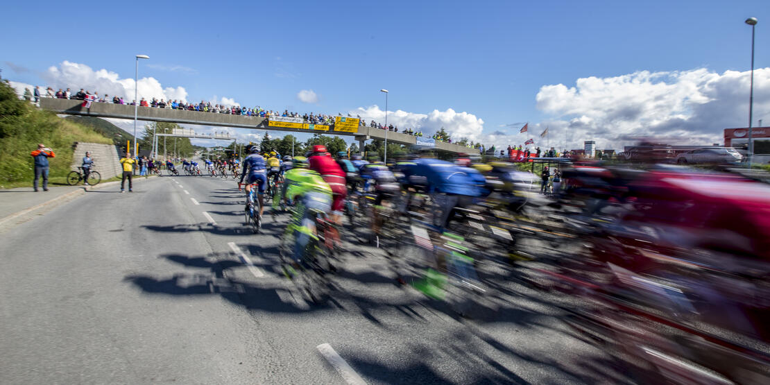 Bilde fra Arctic Race of Norway i Bodø i 2016. Bildet viser syklister i stor fart på riksvei 80. Tilskuere står ved siden av veien og på gangbro over veien. 