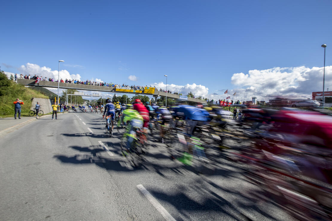 Bilde fra Arctic Race of Norway i Bodø i 2016. Bildet viser syklister i stor fart på riksvei 80. Tilskuere står ved siden av veien og på gangbro over veien . 
