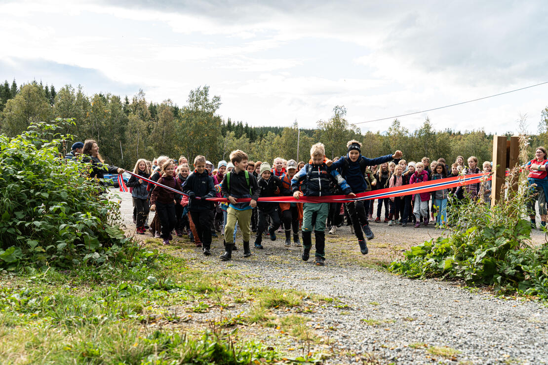 Flere titalls barn løper i uteklær løper mot kamera, og river ned en klippesnor i fargene til det norske flagg. Barna ler mens de løper. I forgrunnen er det litt gress.