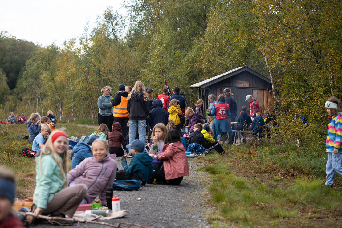 Bildet viser flere titalls elever som sitter på en gruslagt sti og spiser, drikker og slapper av. I bakgrunnen synes noen voksne og en gapahuk. 