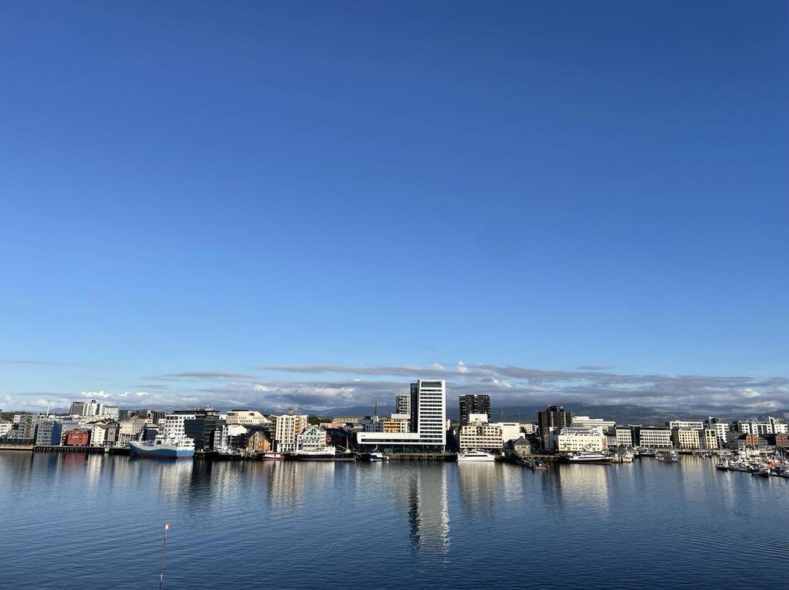 Bildet viser utsikten av skyline til Bodø. I forkant er det hav, mens det lengre inn i bildet er flere hus, små og store, som er vent utover havet.