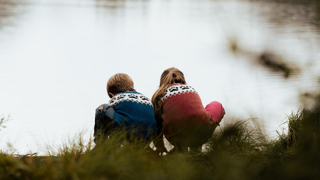 To barn i strikkede gensere sitter på huk med ryggen mot kamera. De sitter på vannkanten og leker med hendene i vannet. 