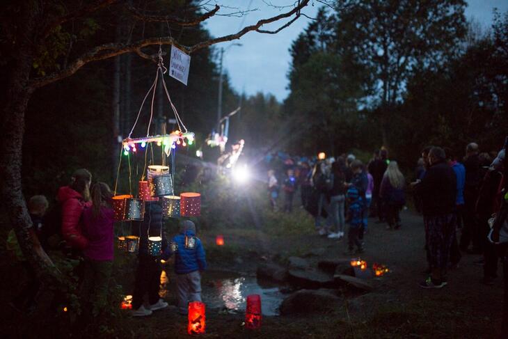 I bakgrunnen, uskarpt, står det mange personer i uteklær. I forgrunnen henger det hjemmelagde lykter fra en grein som er det eneste som er skarpt i bildet. 