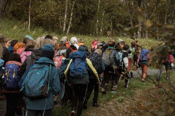 En stor gruppe barn går på en sti i skogen. Barna går med ryggen til og har skolesekker. Trær og gress rundt stien.