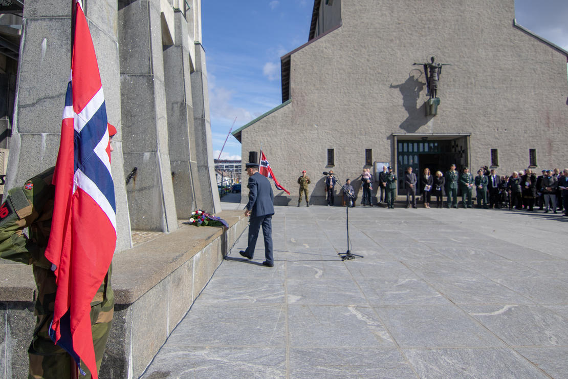 Norsk flagg står ved klokketårnet til Bodø domkirke. Krans er lagt ned ved foten av klokketårnet. Personer står rundt. 
