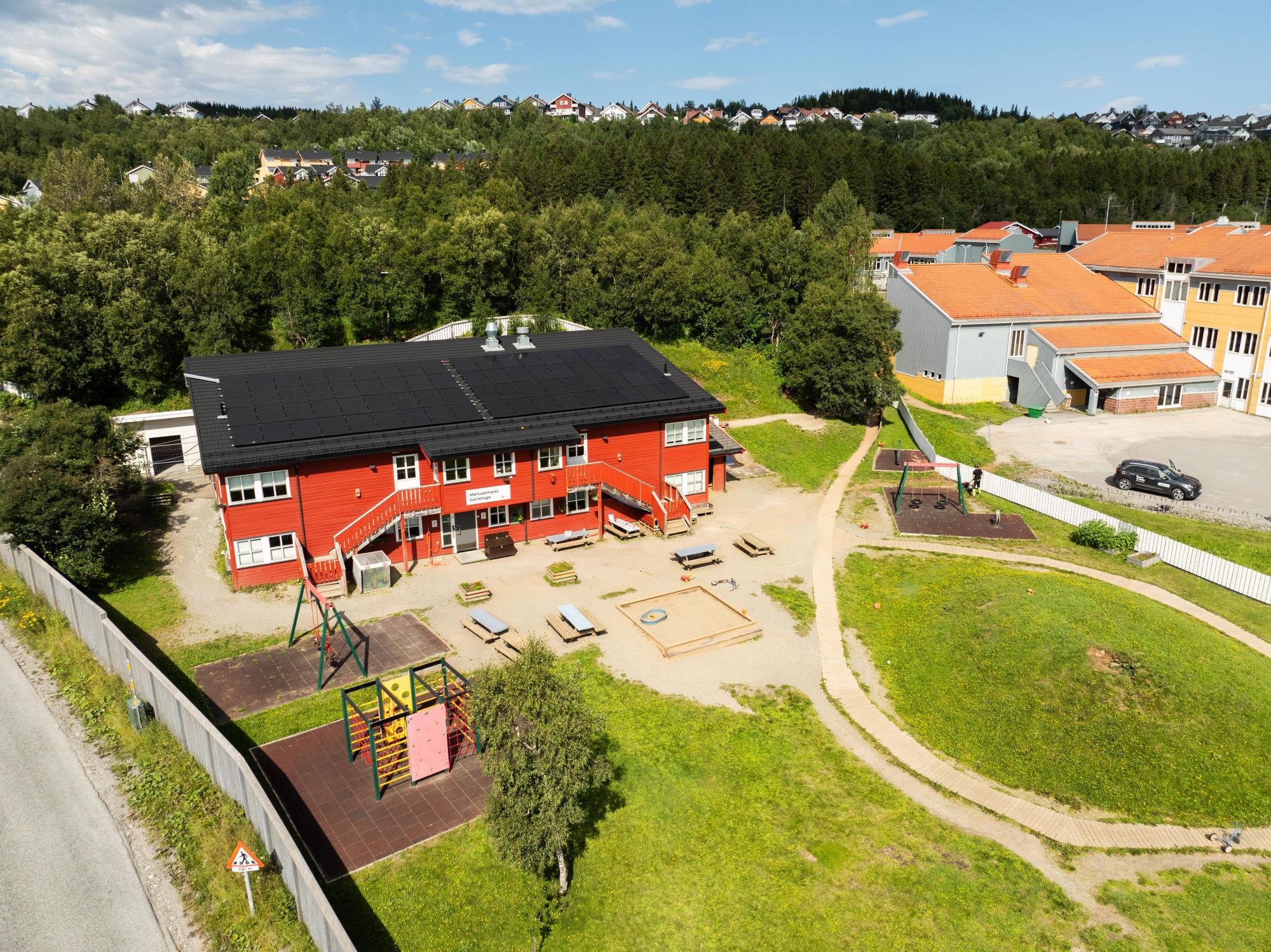 Dronefoto av et rødt stort bygg. Utenfor bygget er det et stort grønnkledd uteområde med lekeapparater og en sandkasse. I bakgrunnen synes skog og blå himmel. - Klikk for stort bilde