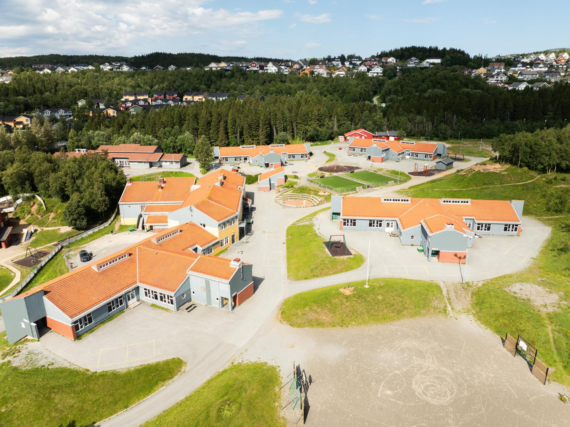 Flere lyseblå bygg med røde steintak står spredt ut på et stort skoleområde. Flere steder er det gressflekker, og i bakgrunnen er det skogsområder med en rekke personlige eiendommer som synes. Bildet er tatt fra luften. - Klikk for stort bilde