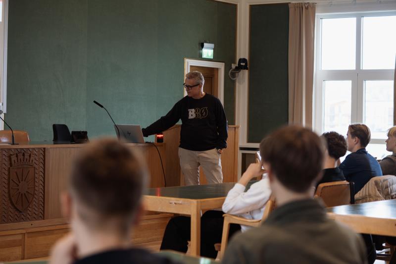 En mann står fremst i et stort rom. Foran han sitter ungdommer og følger med på det han sier. Veggene er grønne og rommet er stort. - Klikk for stort bilde