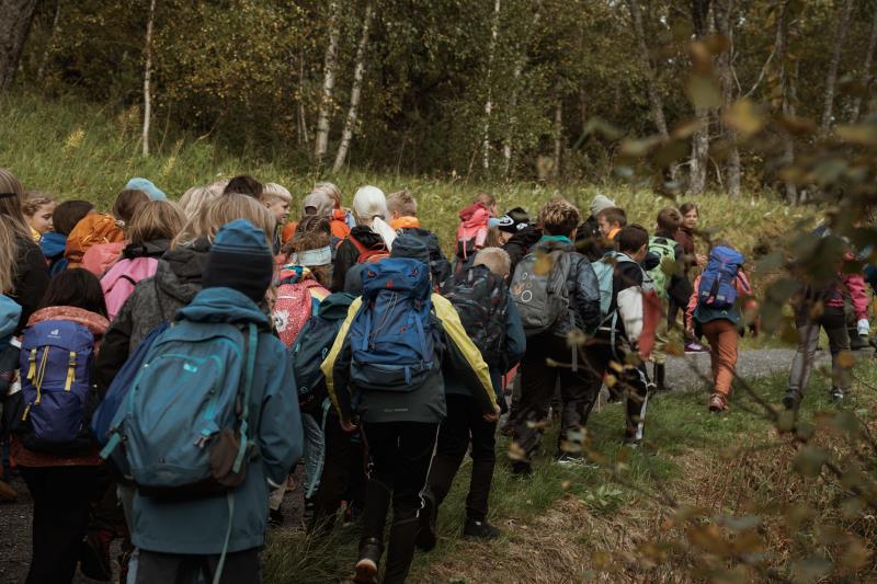 Mange barn med skolesekker på ryggen går på en sti i skogen. Trær rundt stien. - Klikk for stort bilde