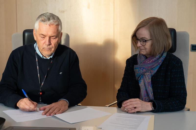 To personer som sitter ved et bor og signerer papirer. - Klikk for stort bilde