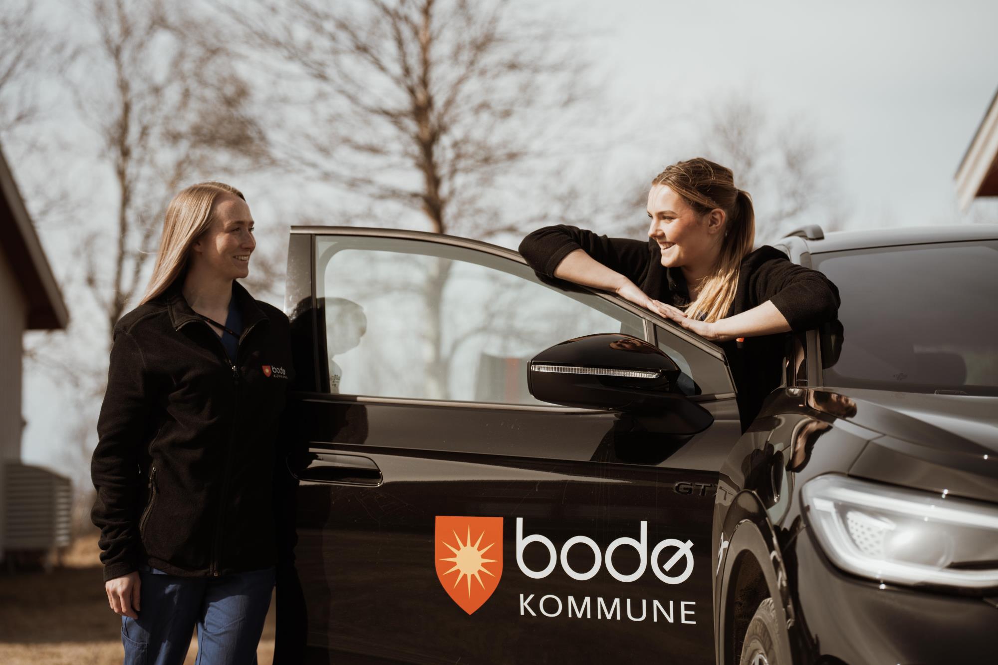  To kvinner står bak døren på en bil mens de ser og smiler til hverandre. Bodø kommunes logo på bildøren. - Klikk for stort bilde