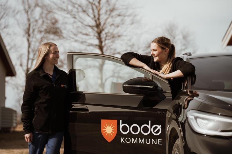  To kvinner står bak døren på en bil mens de ser og smiler til hverandre. Bodø kommunes logo på bildøren. - Klikk for stort bilde
