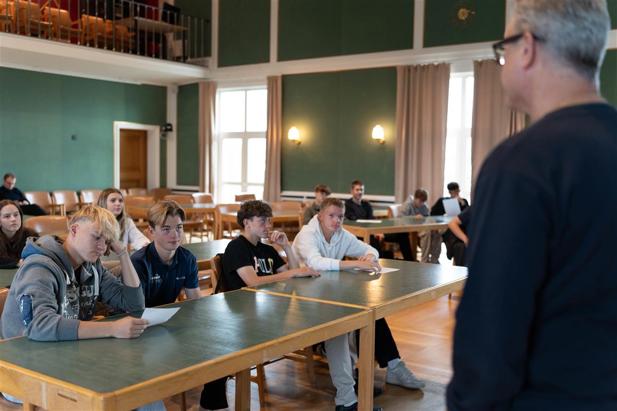 En mann står fremst i et stort rom. Foran han sitter ungdommer og følger med på det han sier. Veggene er grønne og rommet er stort. - Klikk for stort bilde