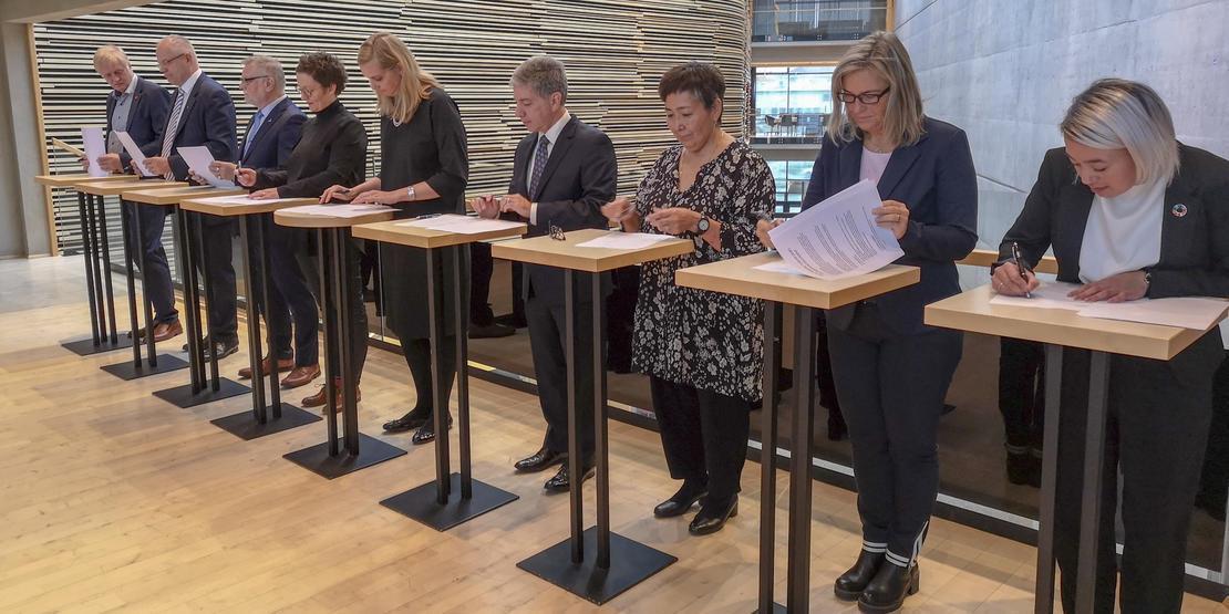 Ni personer står på rekke bak hvert sitt bord og signerer en felles avtale. - Klikk for stort bilde